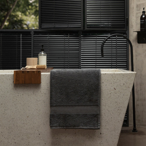 Nebia Bath Towel Gray hanging from a bath tub with other shower accessories and black blinds showing in the background